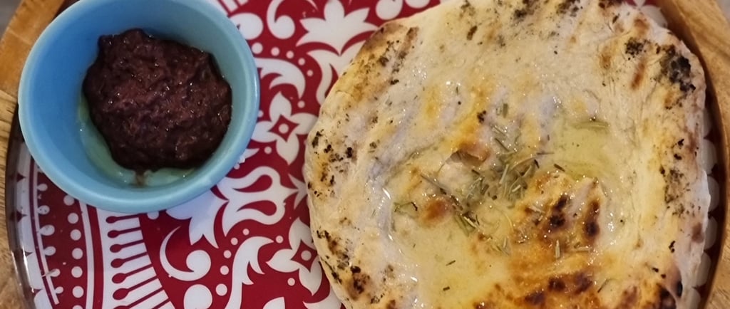 Freshly baked pita bread with olive oil and rosemary served with olive tapenade on a decorative red plate.