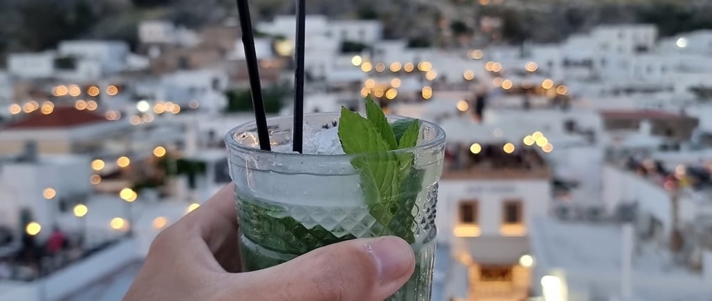 Hand holding a refreshing mojito cocktail overlooking the Lindos Acropolis and village at sunset.