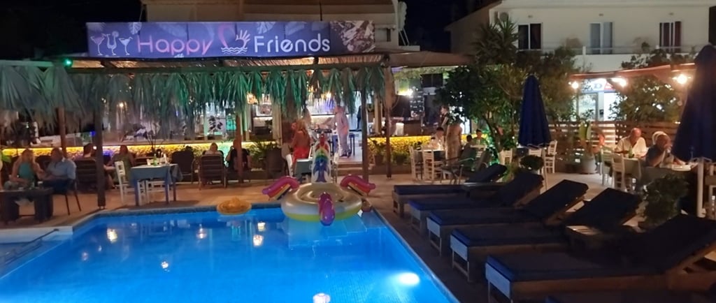 Night view of Happy Friends poolside bar with lit swimming pool and outdoor seating area.