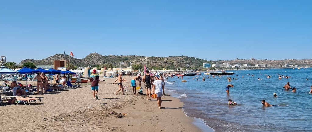 Crowded sandy beach in Rhodes, Greece, with tourists swimming and sunbathing under a clear blue sky.