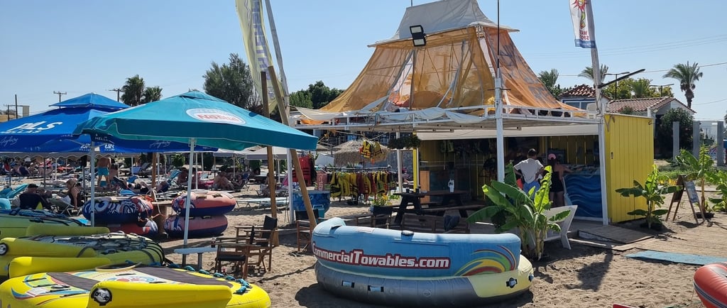 Inflatable water sports tubes and a beach hut bar on a sandy Greek shore under a blue sky.
