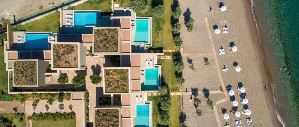 Aerial view of a luxury beachfront resort with private pools and white umbrellas on a sandy shore.