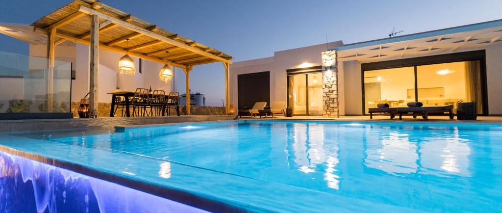 Luxury villa with an illuminated infinity pool and outdoor dining pergola at sunset.