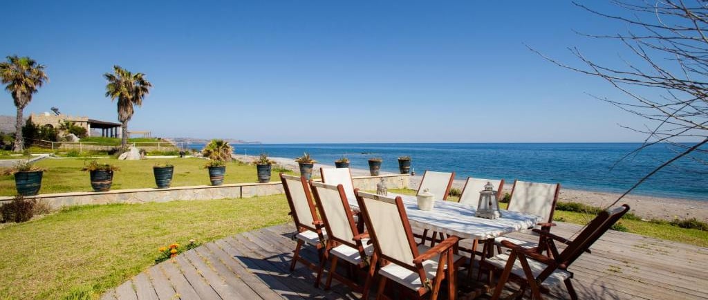 Outdoor dining table on a wooden deck overlooking a beach and clear blue ocean.
