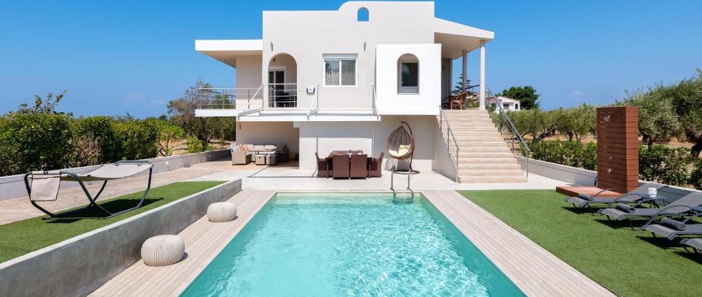 Luxury white villa with a turquoise swimming pool, outdoor dining, and lounge area under a clear blue sky.