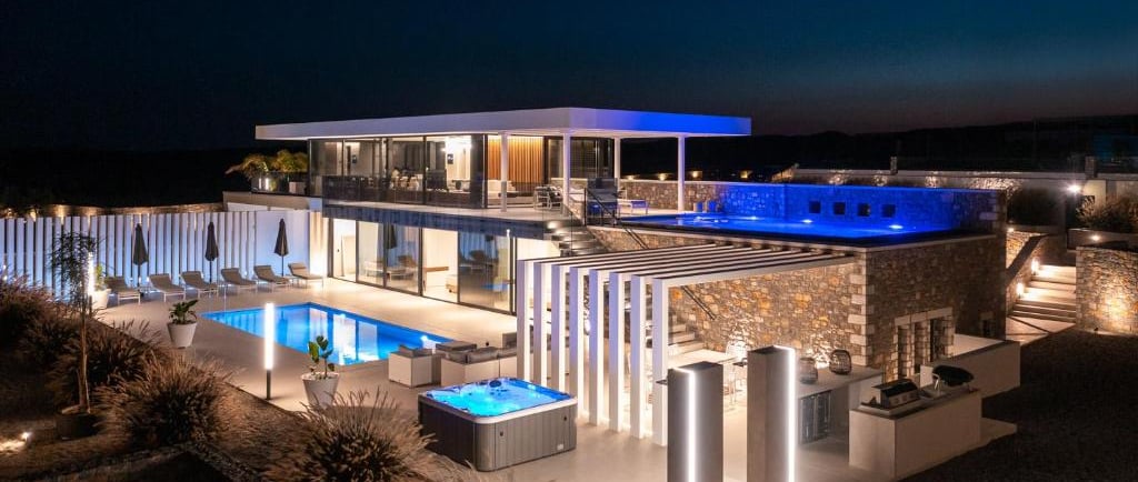 Luxury modern villa at night with illuminated swimming pools, hot tub, and outdoor lounge areas.