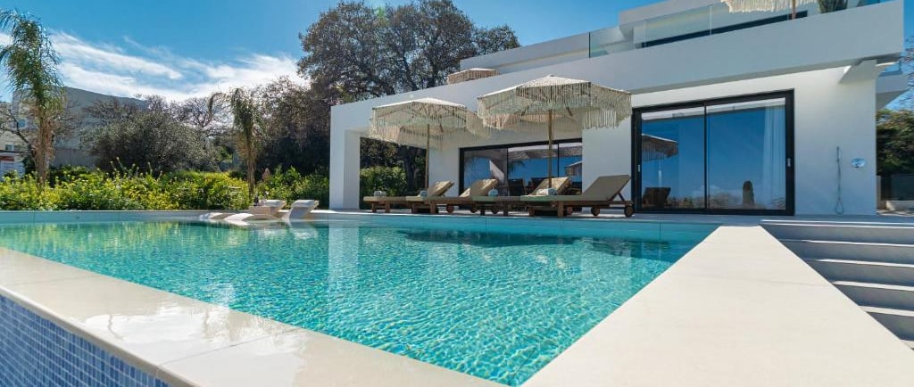 Luxury modern villa with a blue infinity pool, lounge chairs, and sun umbrellas under a clear sky.