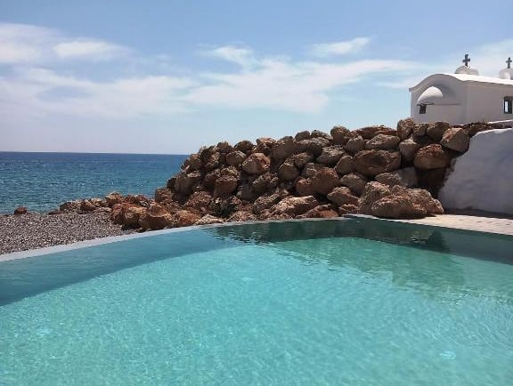 Luxury infinity pool overlooking the Mediterranean Sea with a white Greek chapel and rocky coastline.