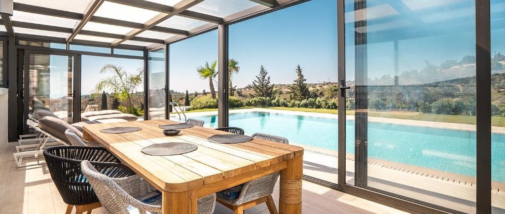 Luxury sunroom with a wooden dining table overlooking a backyard swimming pool and scenic landscape.