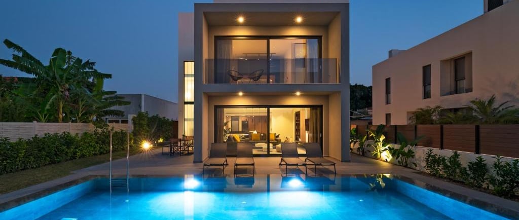 Modern luxury villa at twilight with a glowing blue swimming pool and outdoor lounge area.