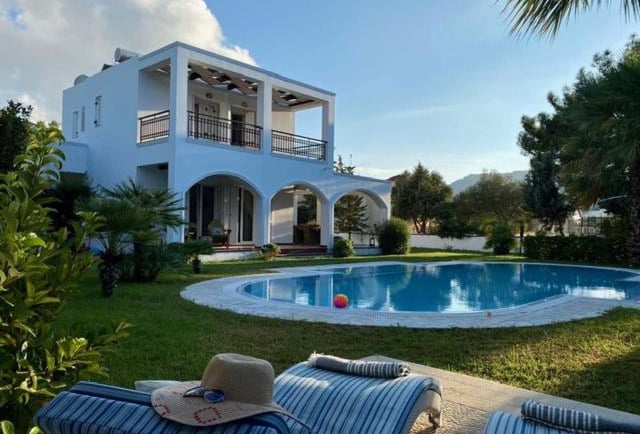 Luxury white villa with private swimming pool, garden, and sun loungers under a blue sky.