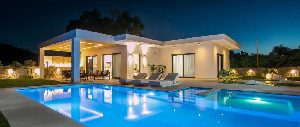Modern luxury villa at night with a glowing swimming pool and outdoor lounge patio area.
