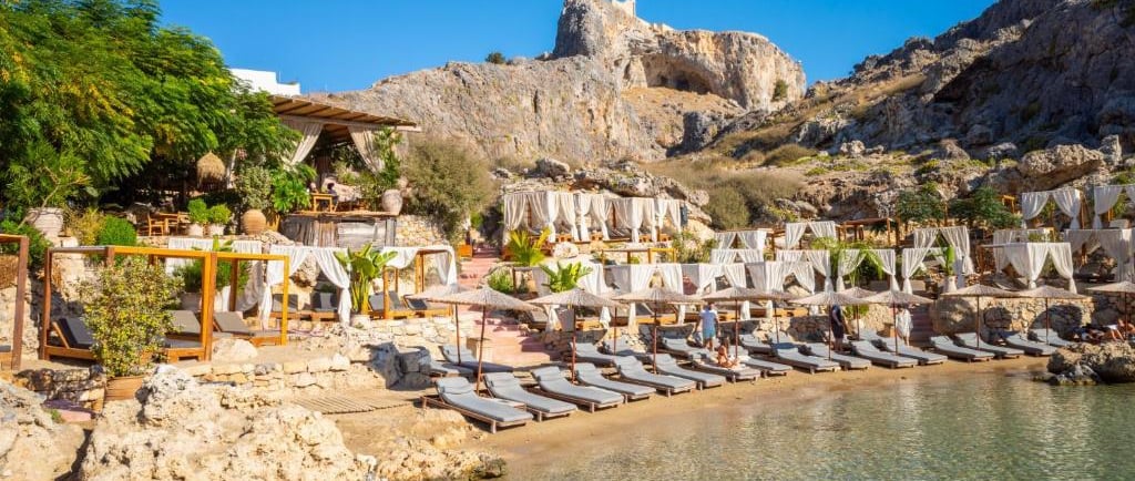Luxury beach club in Lindos, Rhodes, featuring sun loungers and cabanas under the historic Acropolis cliff.