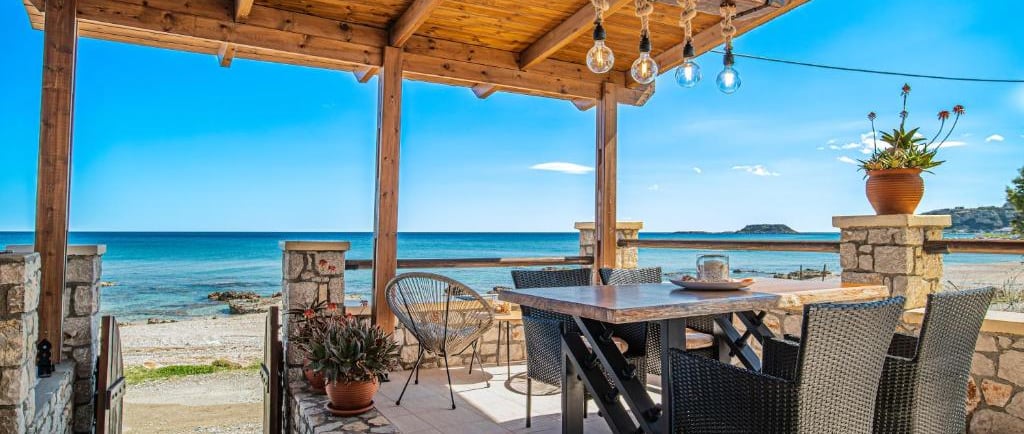 Rustic outdoor patio with dining table and wood pergola overlooking a Mediterranean beach.