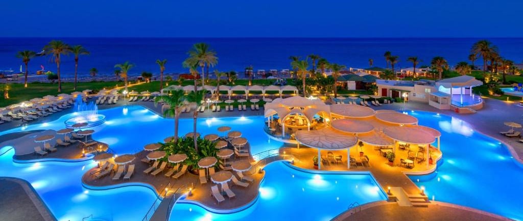 Luxury beach resort at night featuring illuminated swimming pools and palm trees by the ocean.