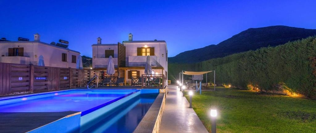 Luxury villa with illuminated private swimming pool and garden at night under a blue sky.