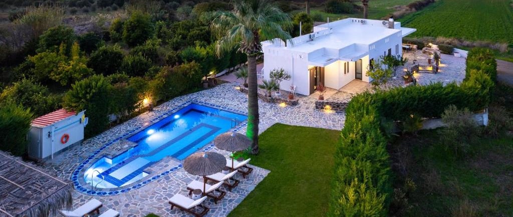 Aerial view of a luxury white villa with a private pool and sun loungers at sunset near the ocean.