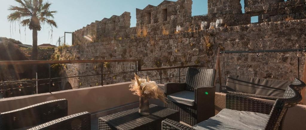 Luxury patio furniture on a sunlit terrace overlooking a historic stone castle wall and palm tree.
