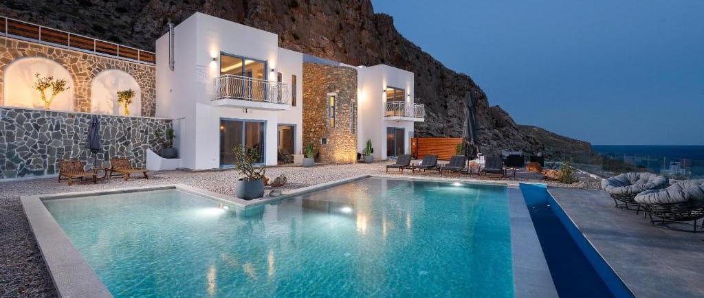 Luxury white villa with illuminated swimming pool at dusk nestled against a rugged mountain cliff.