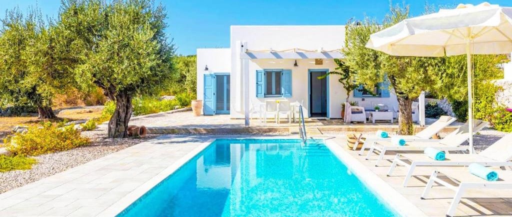 Luxury white Greek villa with a private swimming pool, sun loungers, and olive trees under a clear blue sky.