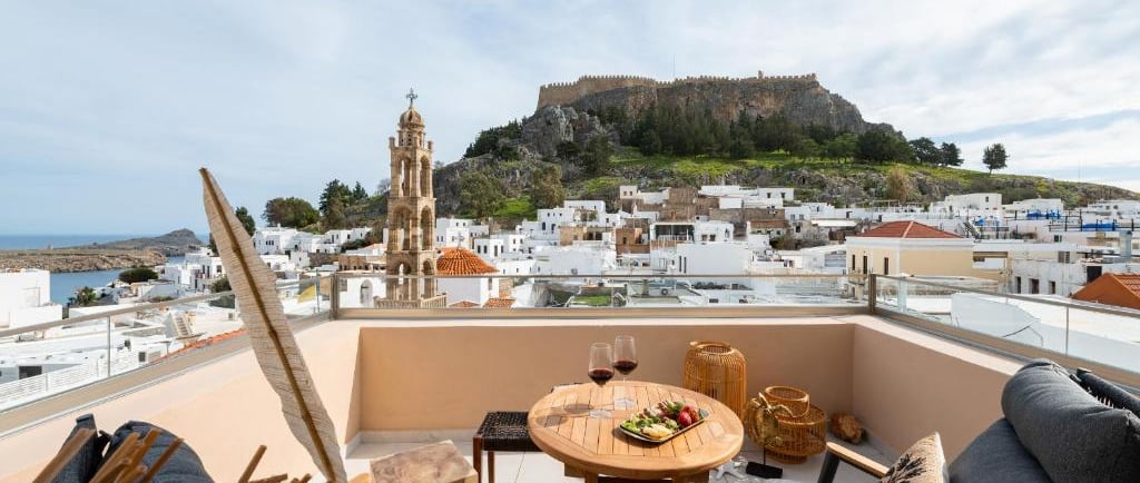 Luxury balcony view of Lindos Acropolis in Rhodes, Greece, with wine and snacks on a patio table.