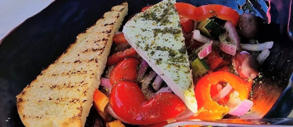 Fresh Greek salad with a wedge of feta cheese, peppers, and grilled bread in a black bowl.