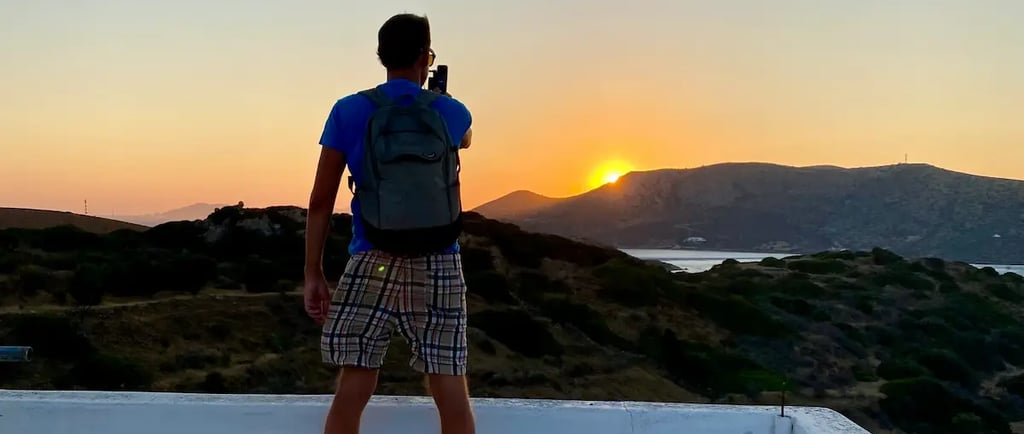 A male traveler with a backpack takes a sunset photo from a rooftop overlooking mountains and the sea.