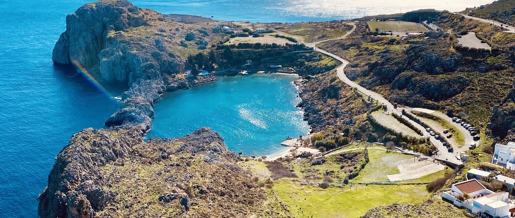 Aerial view of St. Paul's Bay in Lindos, Rhodes, featuring turquoise water, rocky cliffs, and a winding road.