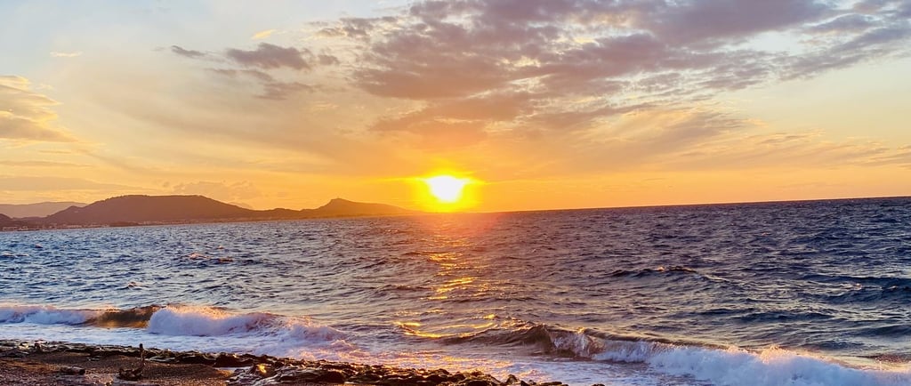 Golden sunset over a rocky pebble beach with ocean waves and a blue cloudy sky.