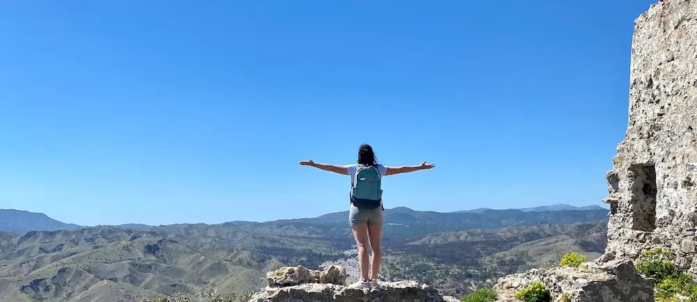 Female hiker with a backpack stands on stone ruins overlooking a vast mountain valley under a clear blue sky.