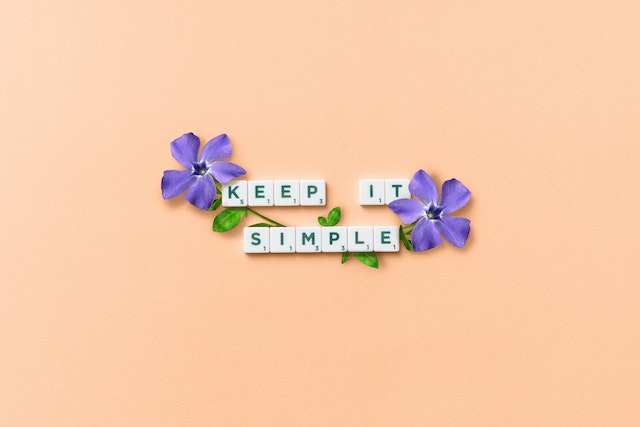 Scrabble tiles spelling out the words keep it simple on an orange background with 2 purple flowers