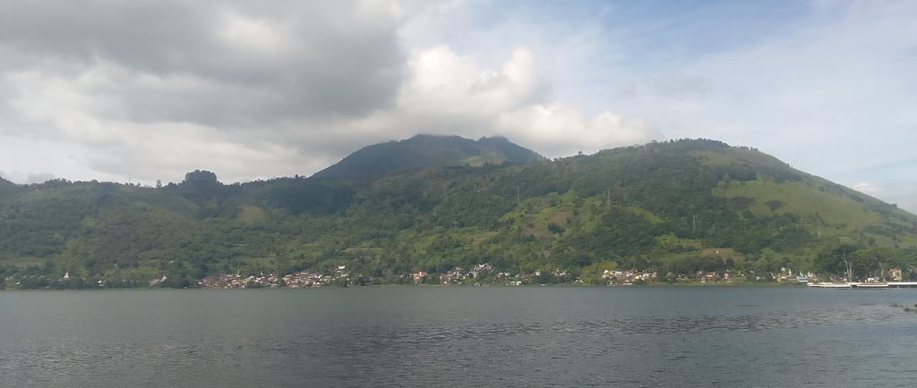 a mountain in the background is getting covered in clouds with houses at the bottom of the mountain by on a lake