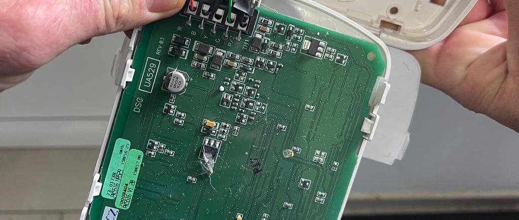 Egy technikus egy riasztórendszer vezérlőpaneljét javítja, amelyen látható a zöld áramköri lap 