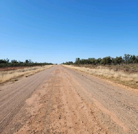 A long, straight dirt road stretching through the remote Australian outback under a clear blue sky.