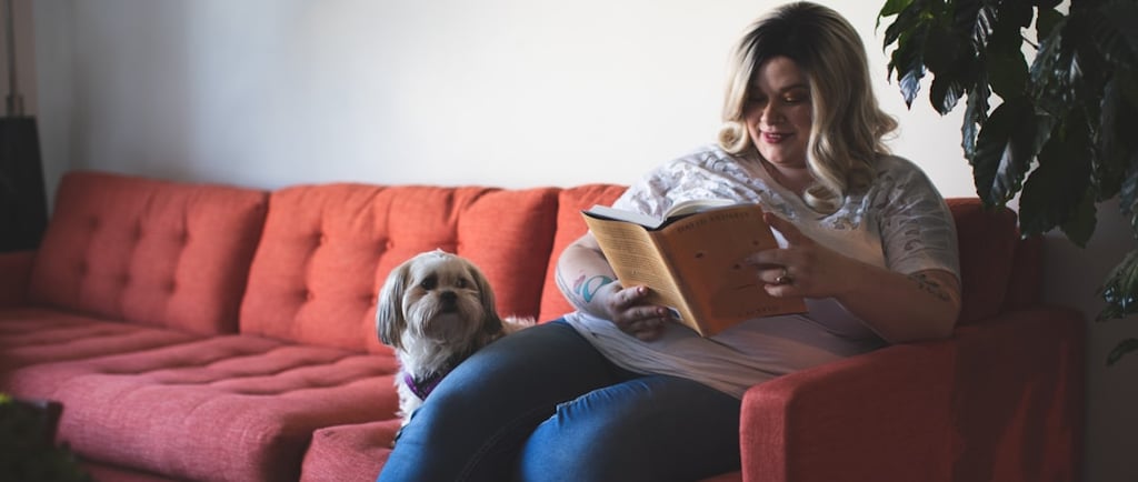 Women sat on a sofa with a dog reading a book.