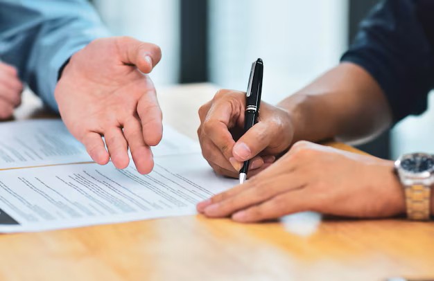 Trata-se de duas mãos, uma assinando um contrato e outra validando a assinatura.