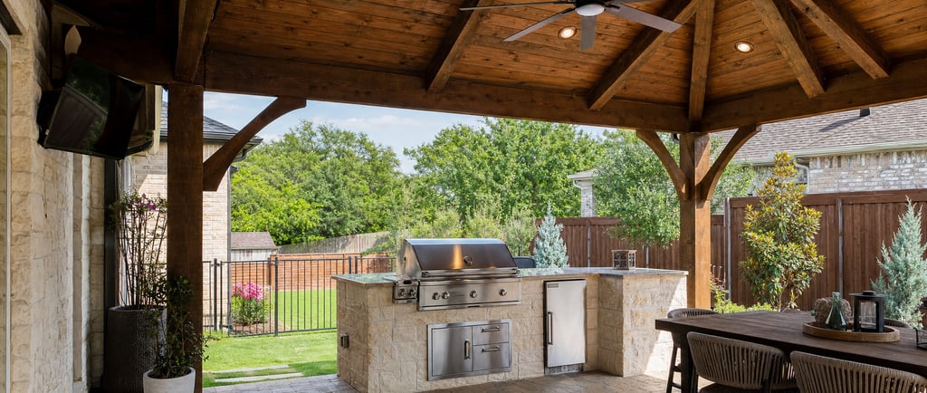 Covered outdoor kitchen in a North Texas backyard with a stone grill island, cedar patio cover, ceiling fan, and shaded seating area