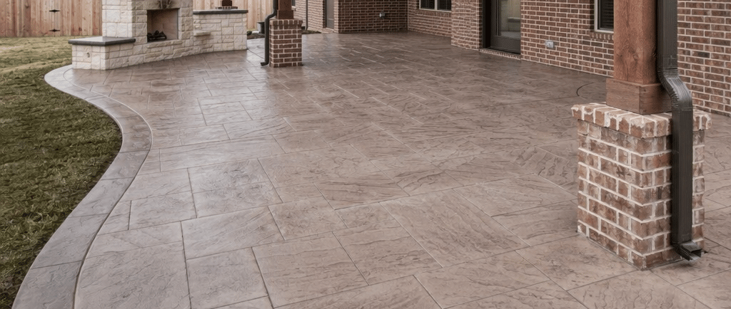 Stamped concrete patio in a fenced brick-home backyard with a light tan stone-pattern finish and a built-in outdoor grill area