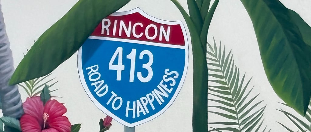 Mural of a Rincon 413 Road to Happiness sign surrounded by tropical hibiscus flowers and banana leaves.