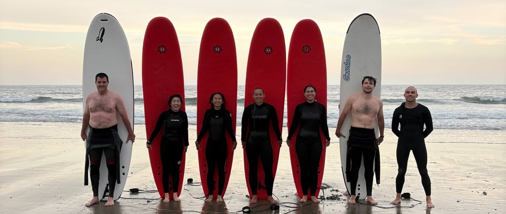 Surfers in wetsuits posing with red and white surfboards on a sandy beach at sunset.