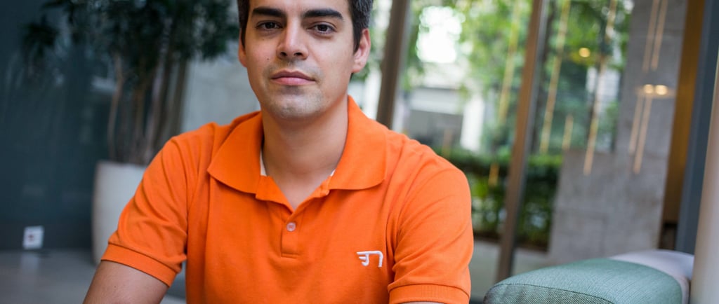 Um homem branco, Tiago Mitraud, veste uma camisa laranja e está sentado em uma cadeira verde.