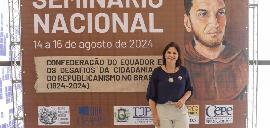 Priscila Krause, mulher branca, em pé na frente de um grande painel, vestindo blusa e calça.