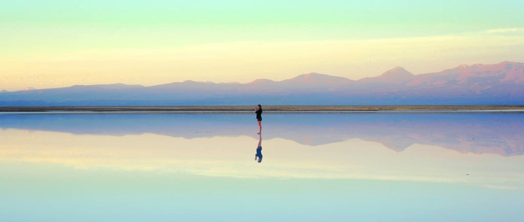 Person standing in a scene of mountains reflected in a lake. Photo by <a href="https://unsplash.com/