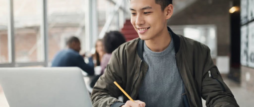 A smiling male student studying on a laptop and taking notes in a modern university library.