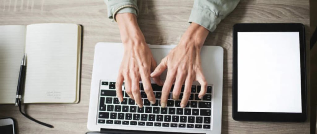Professional person typing on a laptop at a wooden desk with a digital tablet and notebook.