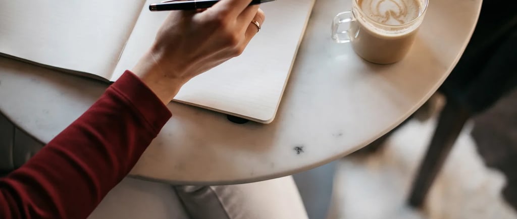A person writing in a blank journal with a pen next to a latte at a cafe table.