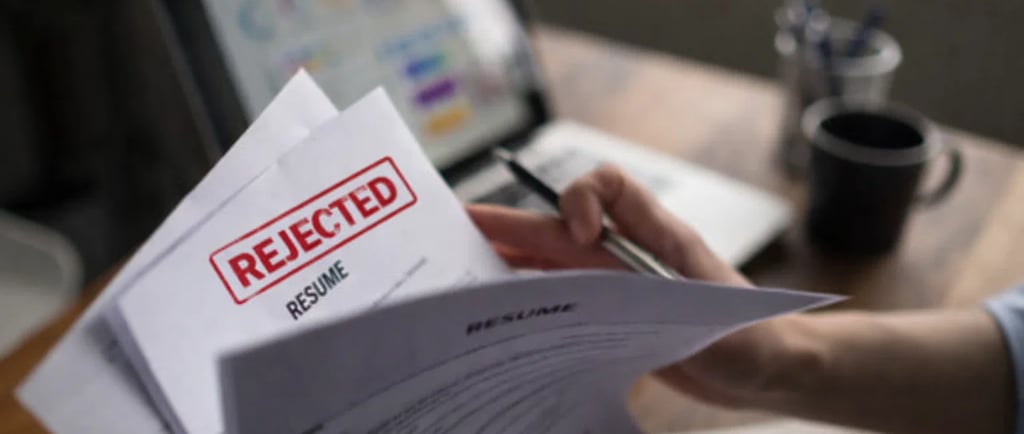 A job applicant holding a resume with a red REJECTED stamp on a wooden desk with a laptop.