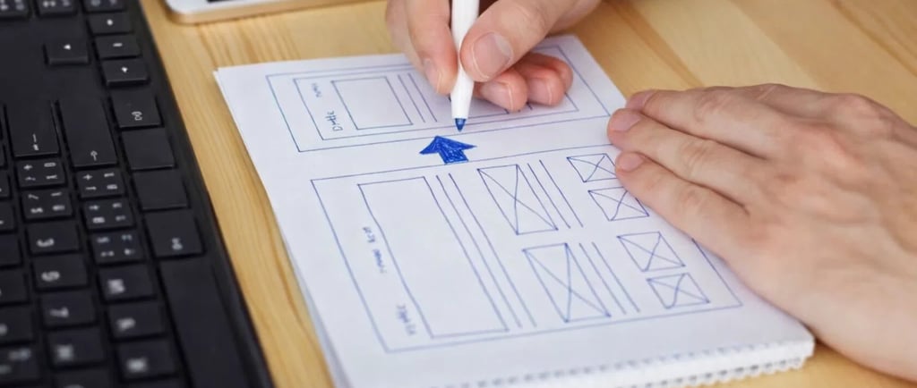 A web designer drawing a UI wireframe sketch on paper at a wooden desk with a computer keyboard.
