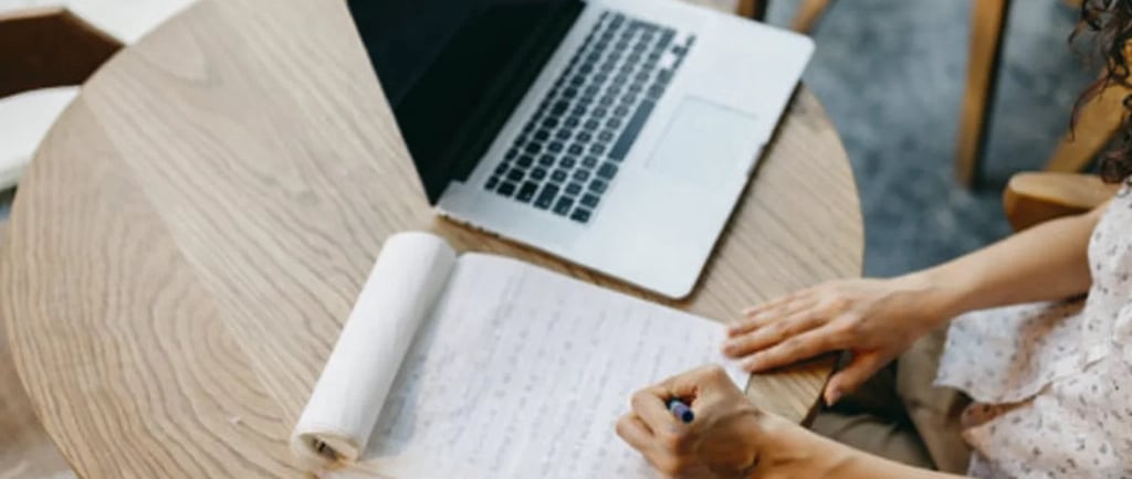 A copywriter taking notes on paper next to an open laptop on a wooden table.