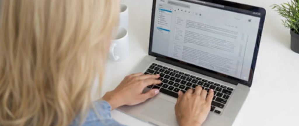 Blonde woman copywriter typing an email on a laptop at a white desk in a bright office.